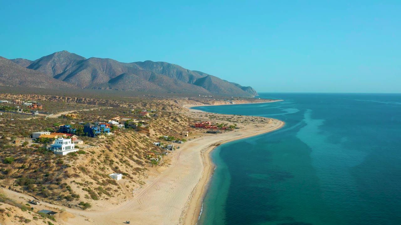 High aerial view in El Sargento La Ventana, Mexico, with turquoise shallows tracing the beach, scattered homes on the dry hillside, distant mountains, and no visible people.