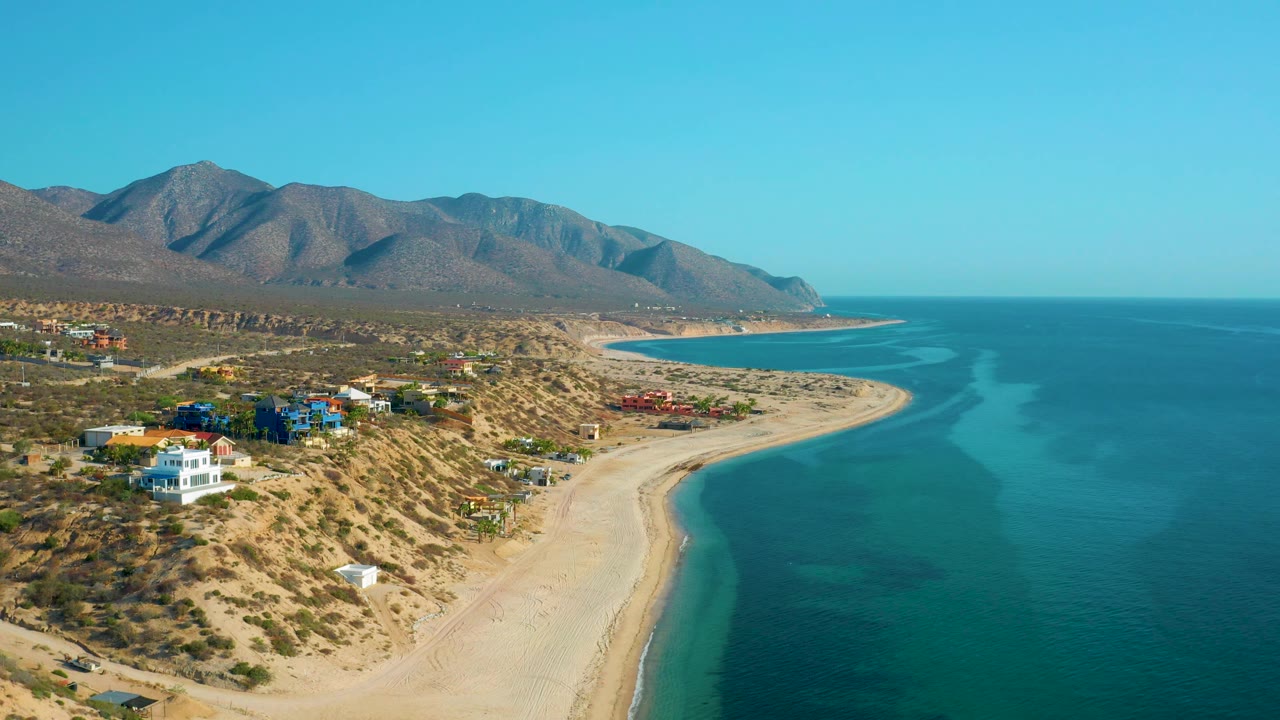 Wide aerial frame from El Sargento La Ventana, Mexico, showing deeper blue water on the right, a pale beach below the hills, colorful hillside houses, mountains, and no visible people.