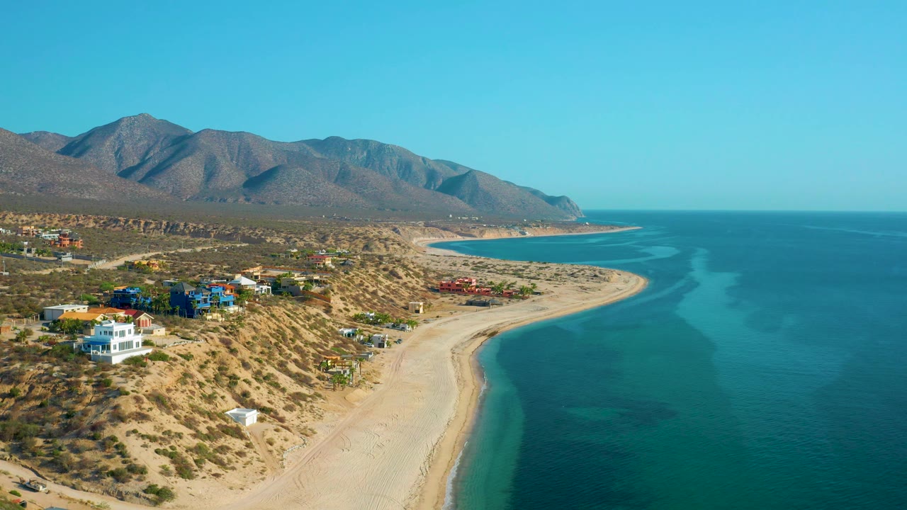 Aerial coastline with broad empty beach, scattered homes, ocean, and mountains.