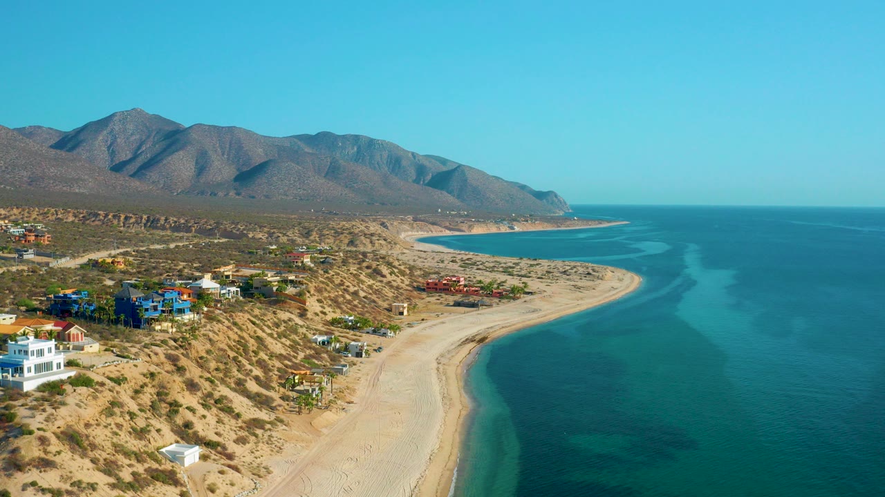Aerial view over El Sargento La Ventana with hillside homes at left, a pale sandy beach curving toward mountains, blue ocean at right, and no visible people.