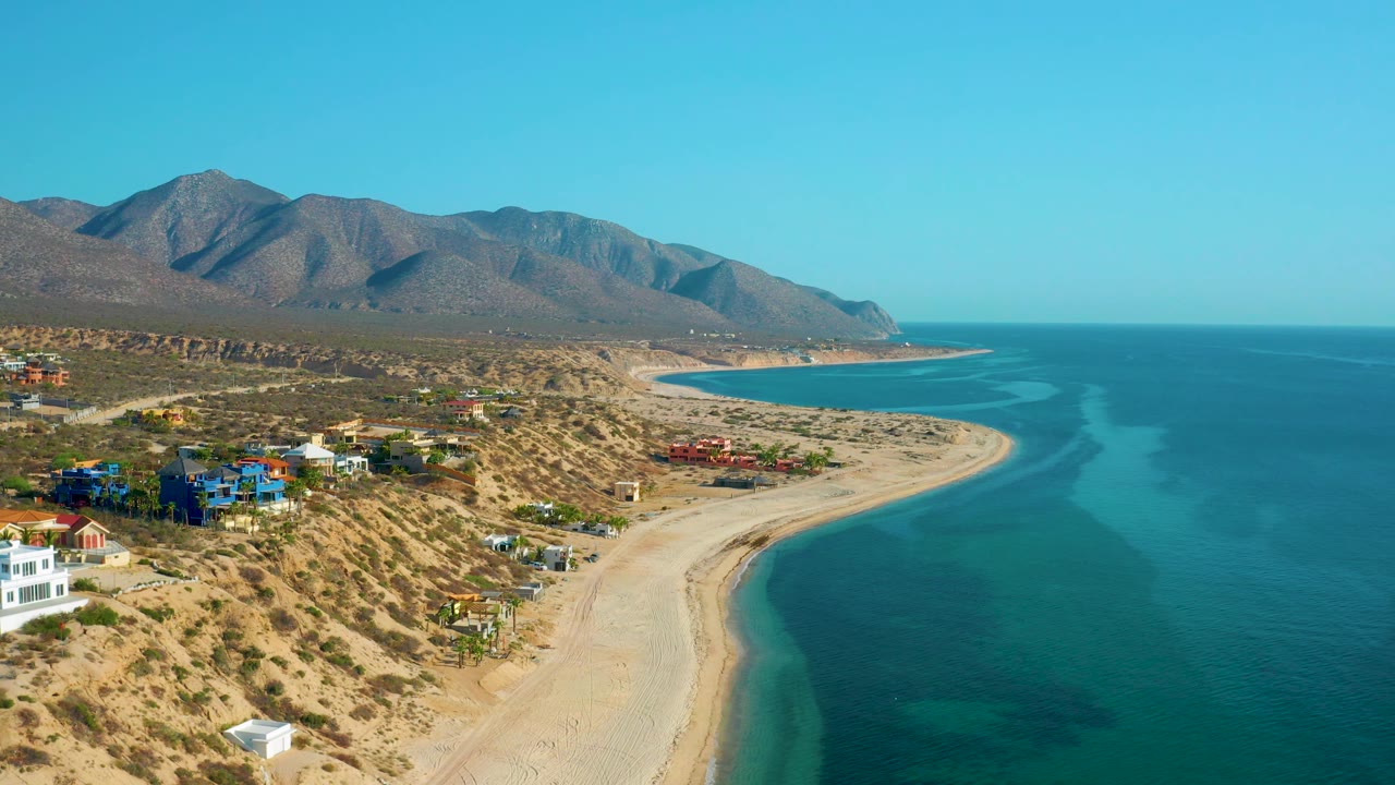 Aerial coastline view in El Sargento La Ventana with bright turquoise shallows near the sand, village homes on the left slope, distant mountains, and no visible people.