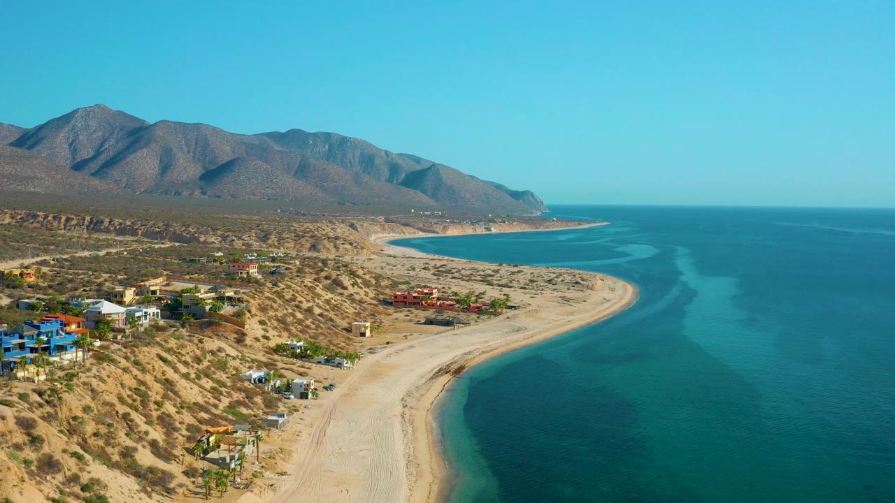 Aerial frame over El Sargento La Ventana, Mexico, showing a curved sandy beach, turquoise water, scattered coastal houses, dry desert terrain, mountains, and no visible people.