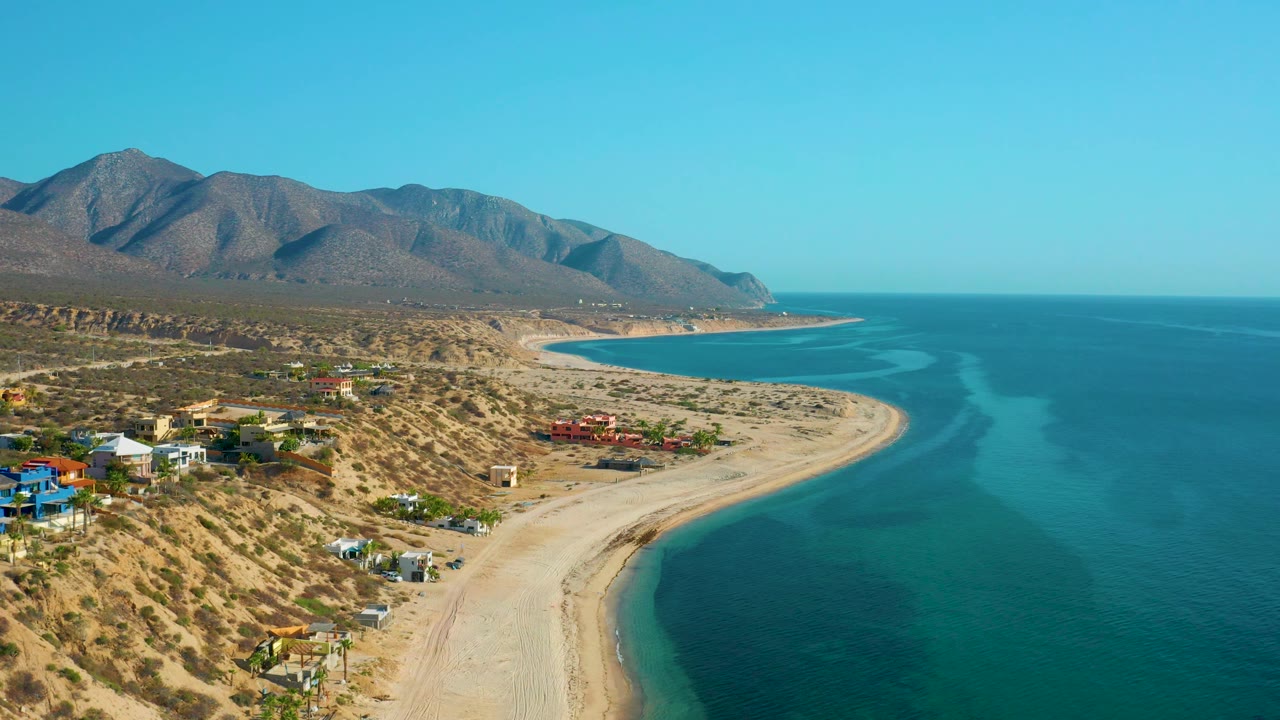 Aerial coastal frame from El Sargento La Ventana, Mexico, showing the beach sweeping around a bay, scattered houses on desert slopes, open ocean, mountains, and no visible people.