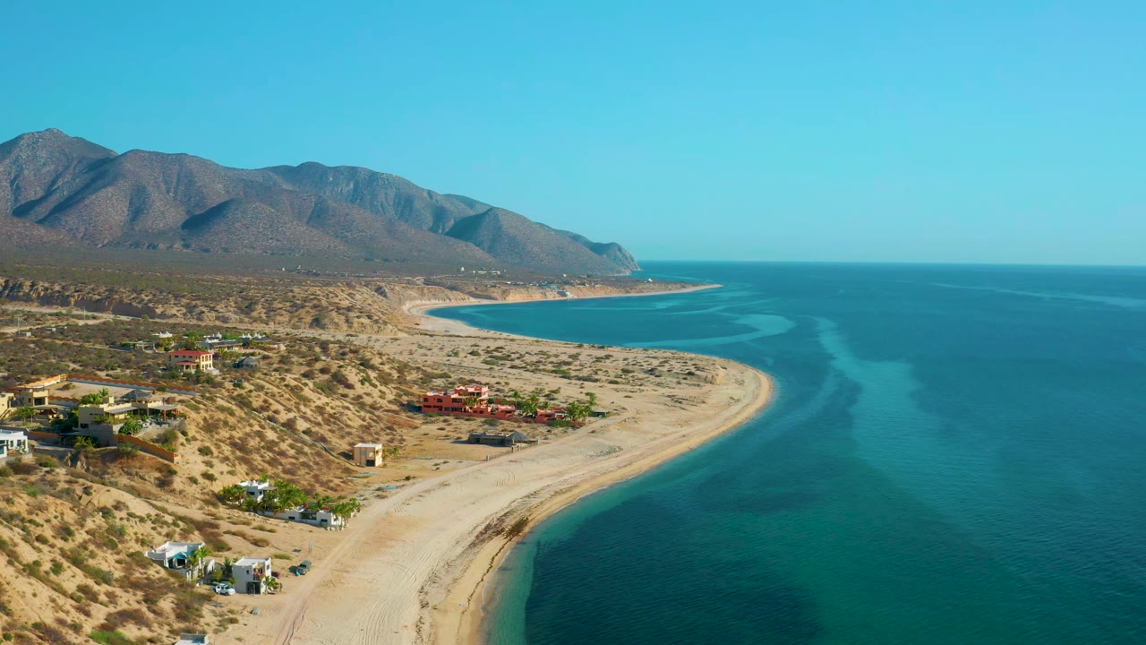 Wide coastal aerial with a sandy shoreline, turquoise water bands, houses, and mountains.