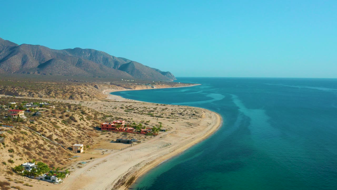 Opening aerial frame over El Sargento La Ventana, Mexico, with a curved sandy shoreline, turquoise water, desert hills, scattered buildings, and no visible people.