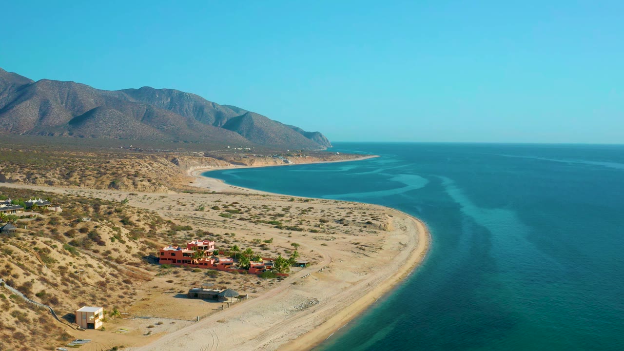 Drone view of a curved sandy shoreline beside blue ocean and desert mountains.