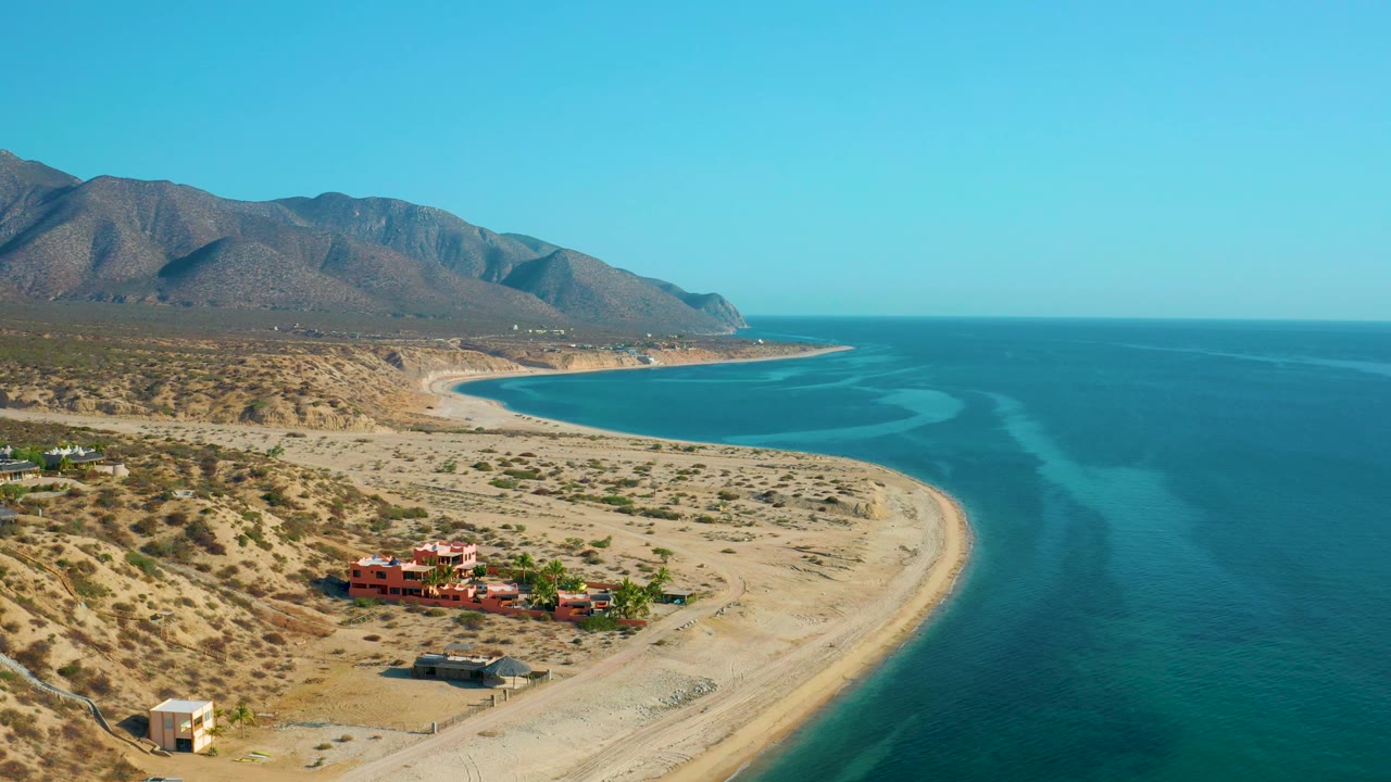 El Sargento La Ventana aerial view with resort-style buildings near the beach, empty sand along the waterline, turquoise sea to the right, mountains to the left, and no visible people.