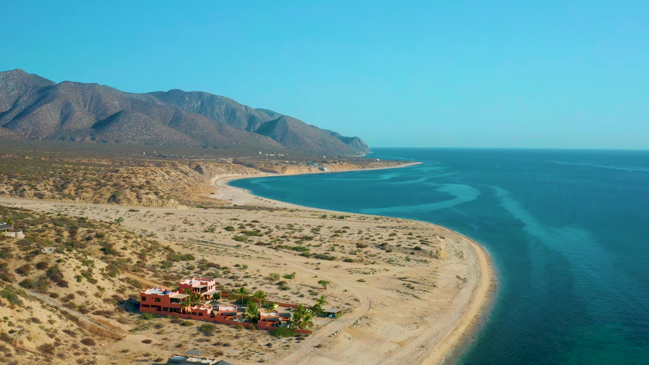 Second aerial frame of El Sargento La Ventana, Mexico, with the red-orange buildings lower in frame, a pale beach curving around the bay, blue ocean to the right, mountains behind, and no visible people.