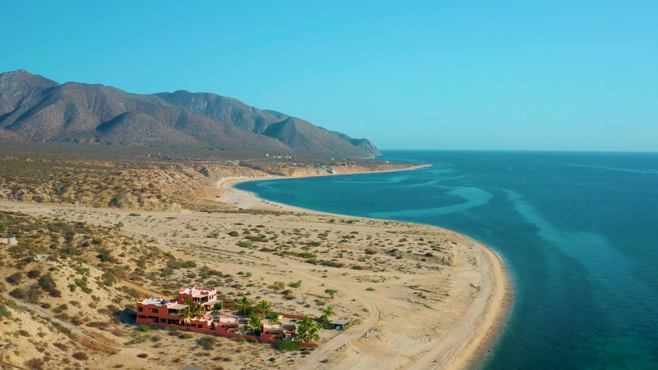 Aerial frame over El Sargento La Ventana, Mexico, where the curved beach and blue water dominate the right side, with dry coastal terrain, red-orange buildings near the lower edge, mountains, and no visible people.