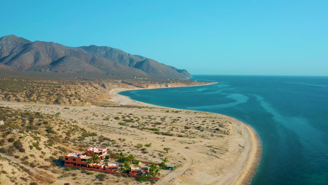 Aerial view with red-orange buildings near the bottom, sandy shoreline, blue sea, scrub hills, and mountains.