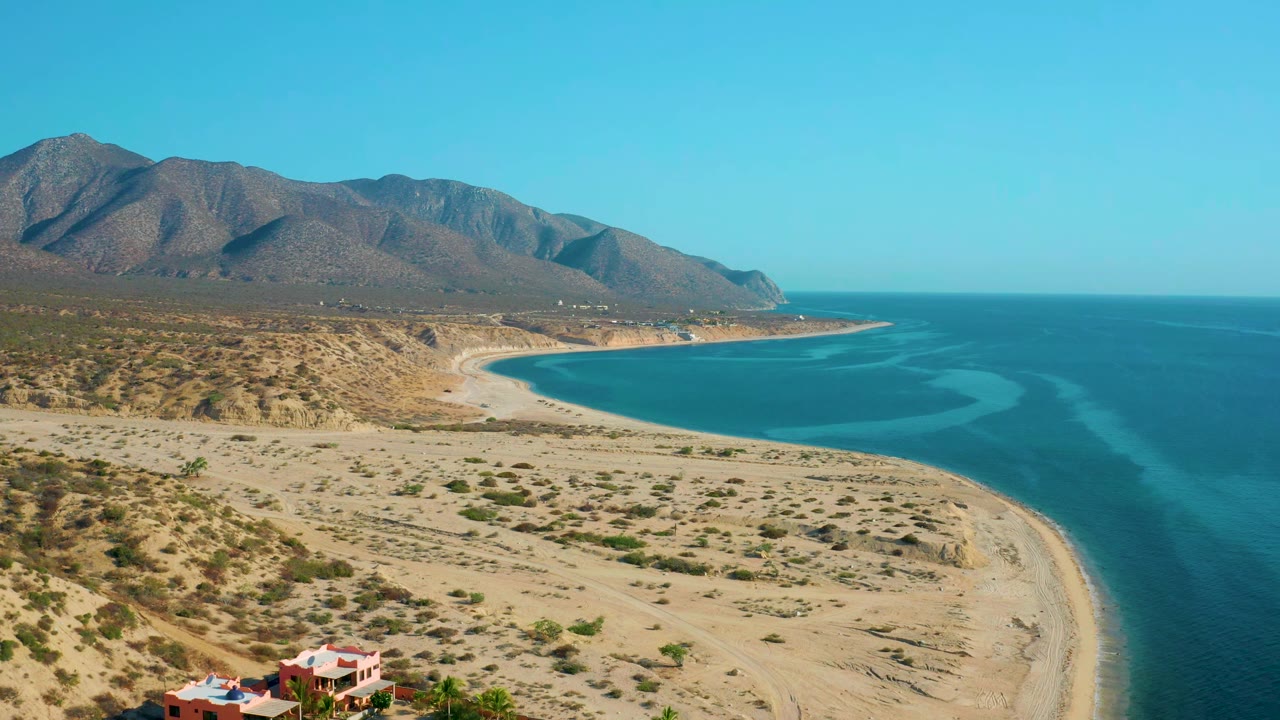 Wide aerial coastline with sandy desert foreground and mountains beyond the bay.