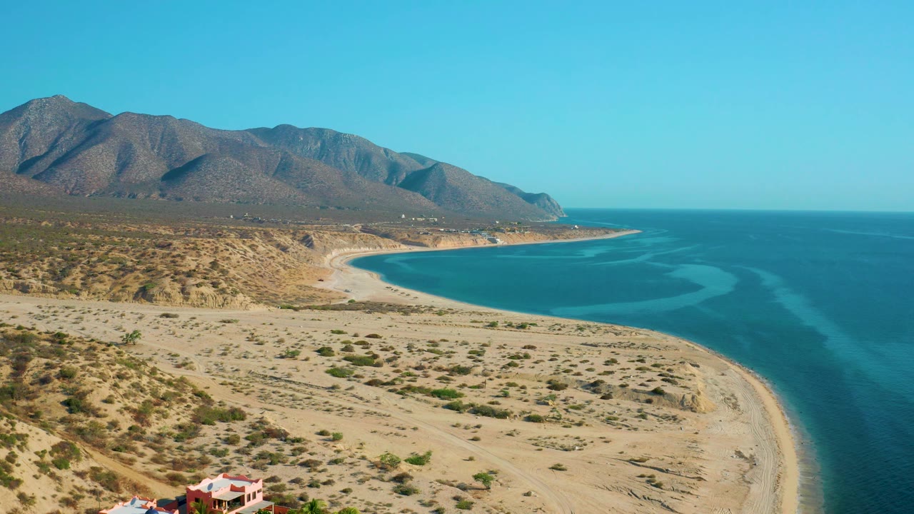 High aerial frame of El Sargento La Ventana with a pale beach arc, open desert flats, blue sea bands, mountains on the horizon, and no visible people.