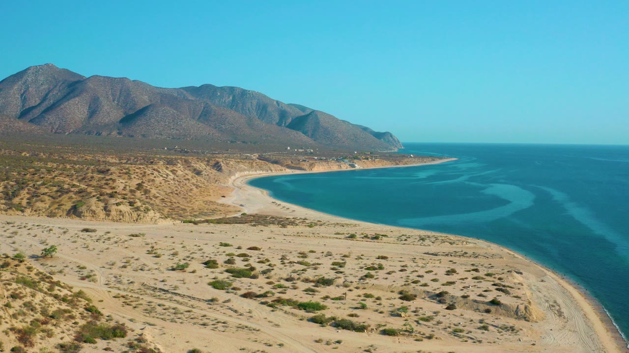 Wide aerial view of desert coast, curved bay, blue ocean, and mountains.