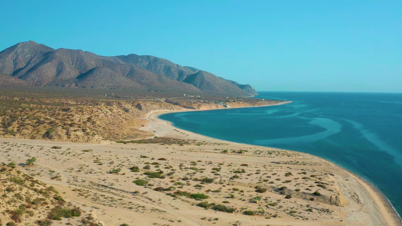 Aerial frame over El Sargento La Ventana, Mexico, showing more sandy foreground and low shrubs before the curved bay, with mountains behind and no visible people.