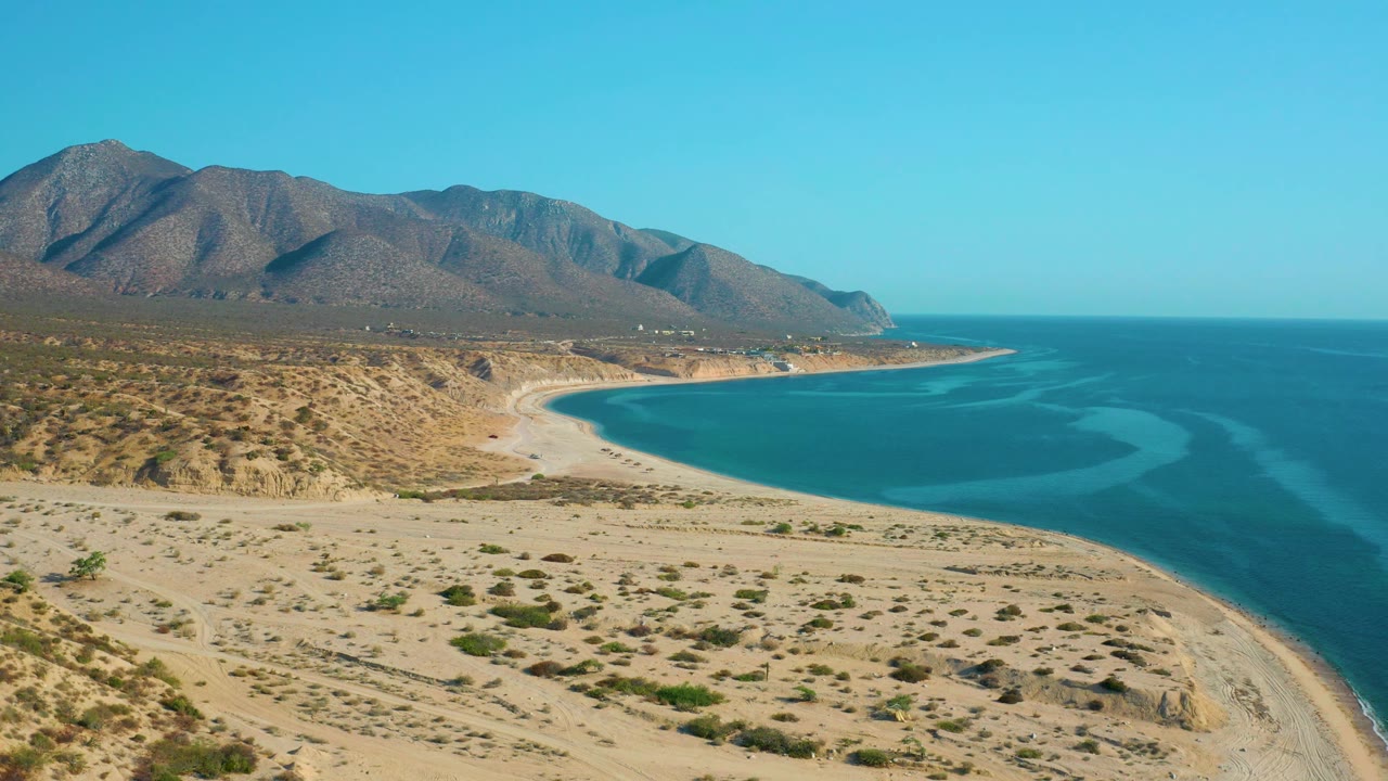High view of El Sargento La Ventana, Mexico, where a pale beach arcs around turquoise water below a long brown mountain range, with no visible people.