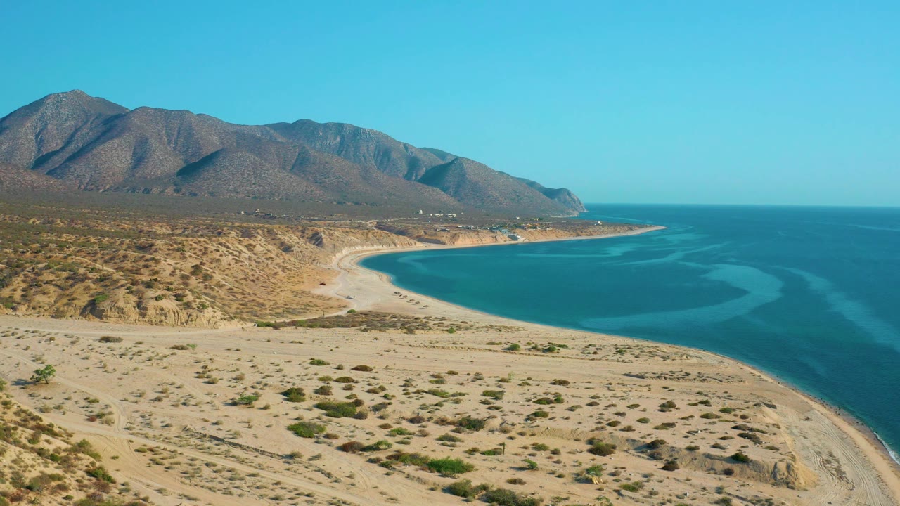 Mid-clip aerial view of El Sargento La Ventana, Mexico, with the mountain range occupying the left background and the ocean filling the right side, with no visible people.
