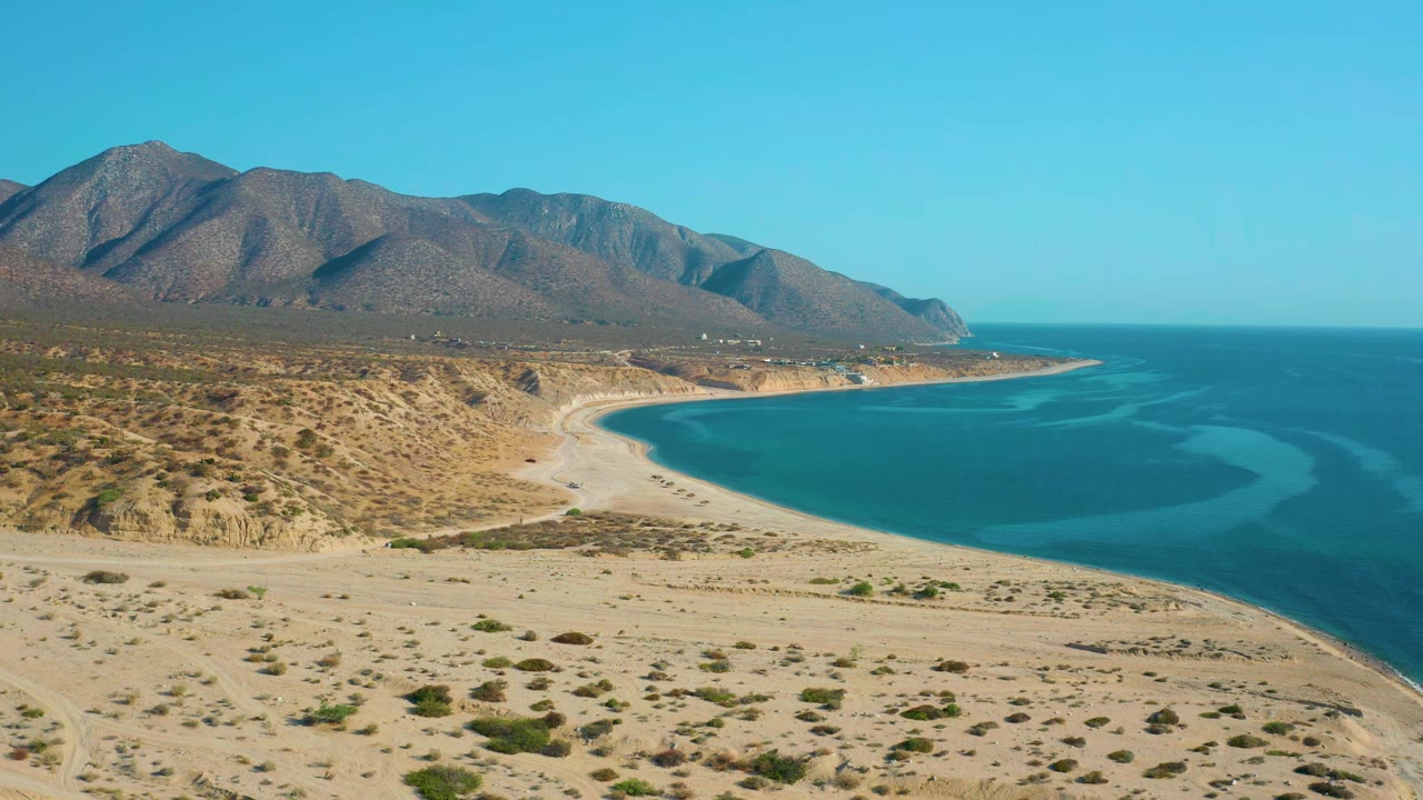 Wide aerial frame over El Sargento La Ventana, Mexico showing the bay's curved shoreline, pale sand, scattered desert brush, blue water bands, distant mountains, and no visible people.