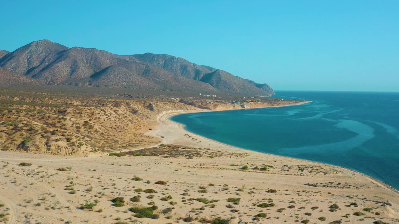 Aerial view in El Sargento La Ventana, Mexico with more open water visible on the right, desert slopes on the left, a pale beach arc below, distant ridges, and no visible people.