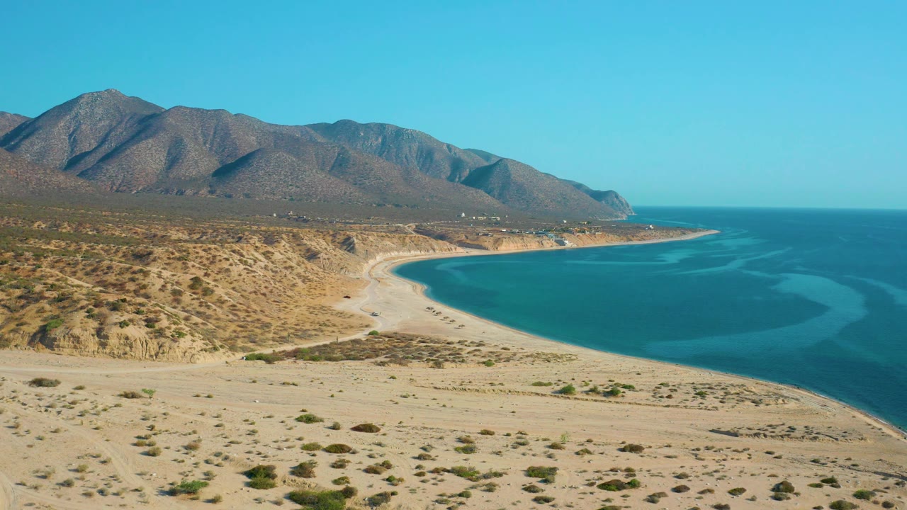 Mid-clip aerial frame over El Sargento La Ventana, Mexico with sandy desert foreground, sparse shrubs, a bright beach curve, calm-looking blue water, mountains, and no visible people.