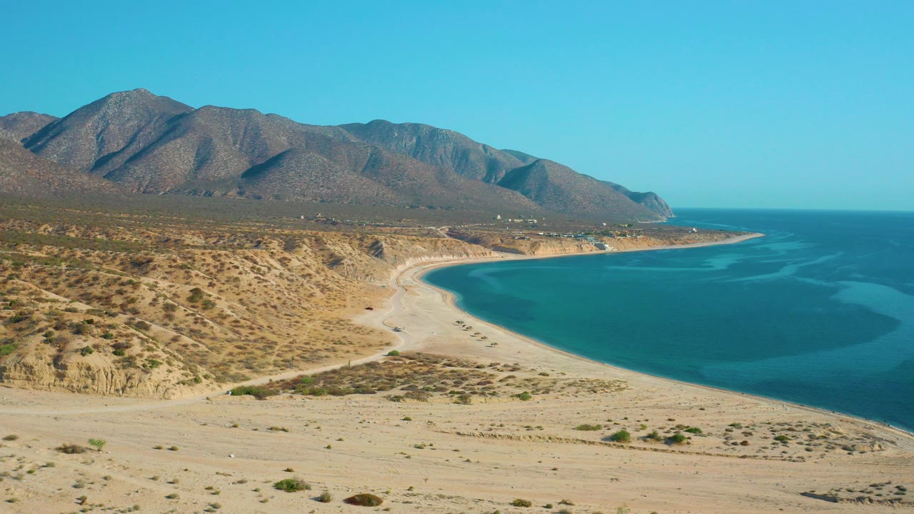 Opening aerial frame over El Sargento La Ventana, Mexico, showing tan desert scrub, a pale beach curve, blue water, distant mountains, and no visible people.