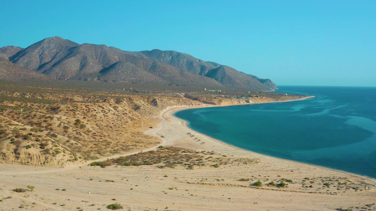Wide aerial frame in El Sargento La Ventana, Mexico, showing the beach arc below rocky mountains, darker blue water offshore, pale sand flats, and no visible people.
