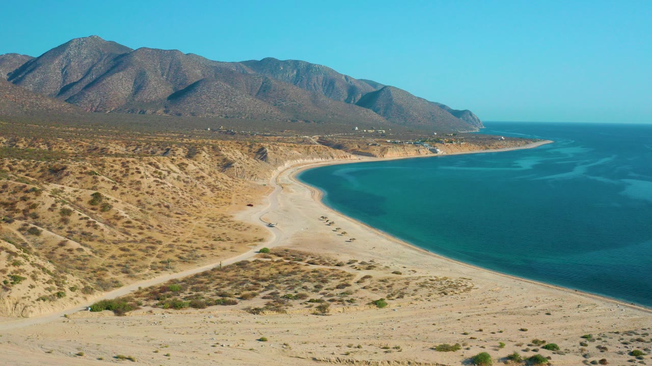 Aerial frame over El Sargento La Ventana, Mexico showing a curving sandy beach beside blue water, scrub-covered terrain, distant mountains, and no visible people.