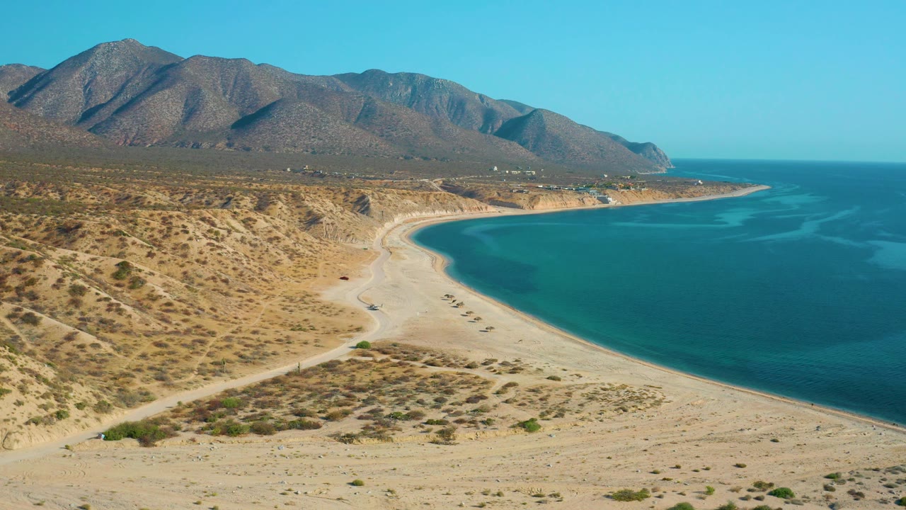 Wide aerial frame over El Sargento La Ventana, Mexico showing desert scrub across the left foreground, a long sandy shore, turquoise water to the right, mountains behind, and no visible people.