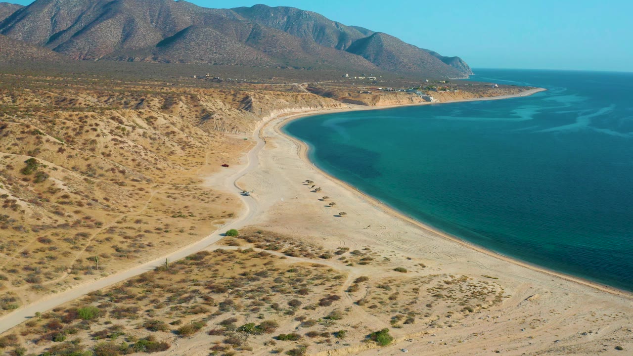 Wide aerial view in El Sargento La Ventana, Mexico, with scrubby desert terrain leading to a sandy beach, turquoise sea on the right, mountains beyond, and no visible people.