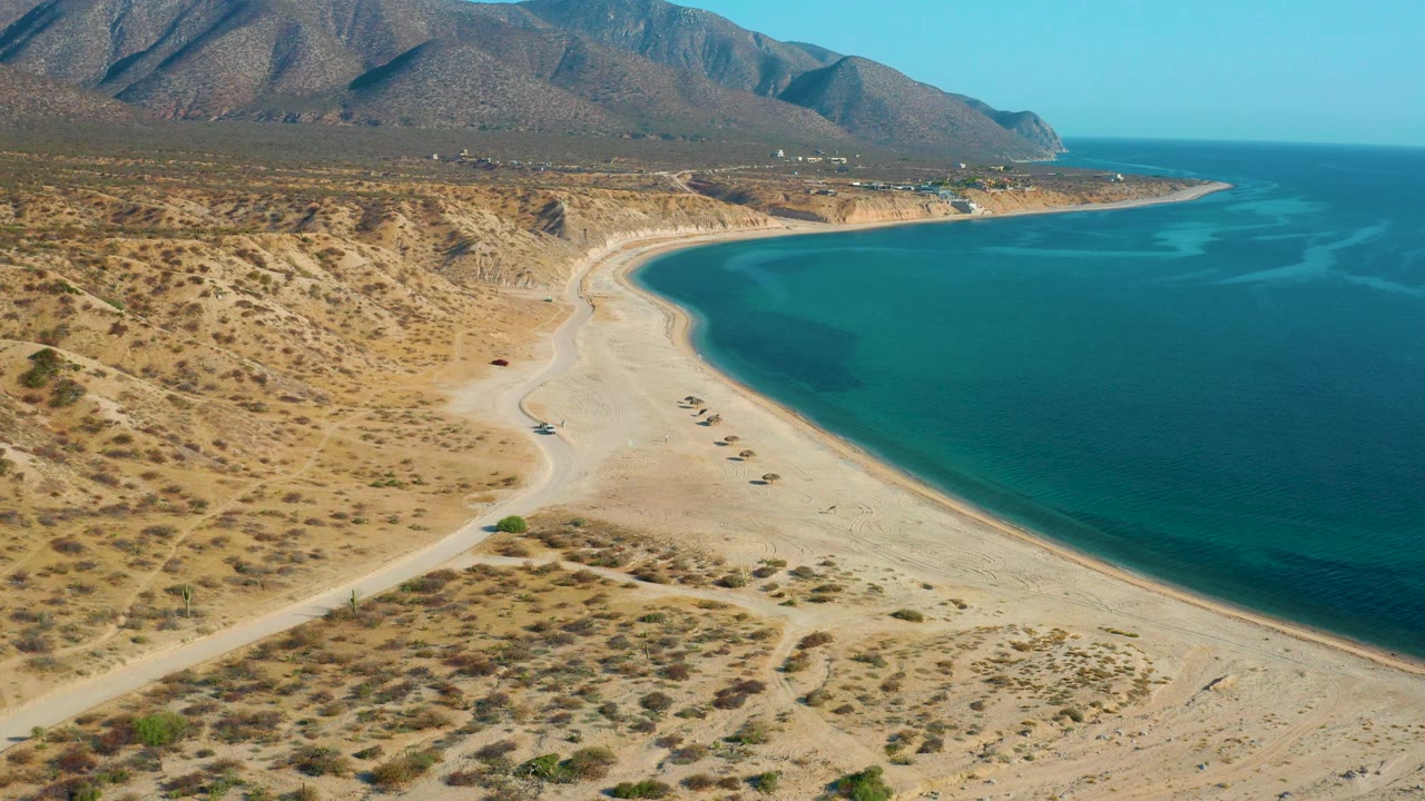 Aerial frame over El Sargento La Ventana, Mexico, showing the beach curve stretching toward a distant point, darker blue water to the right, a mountain ridge above, and no visible people.