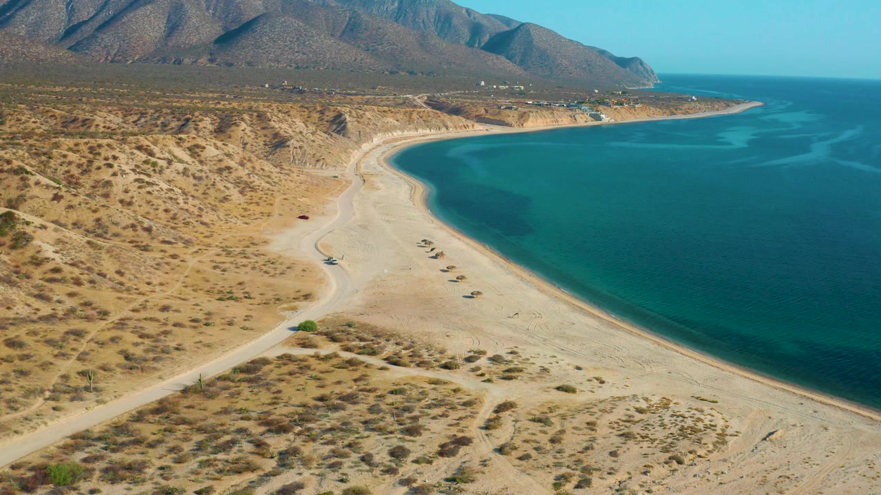 High view of beach access road, sandy shore, blue sea, and arid hills.