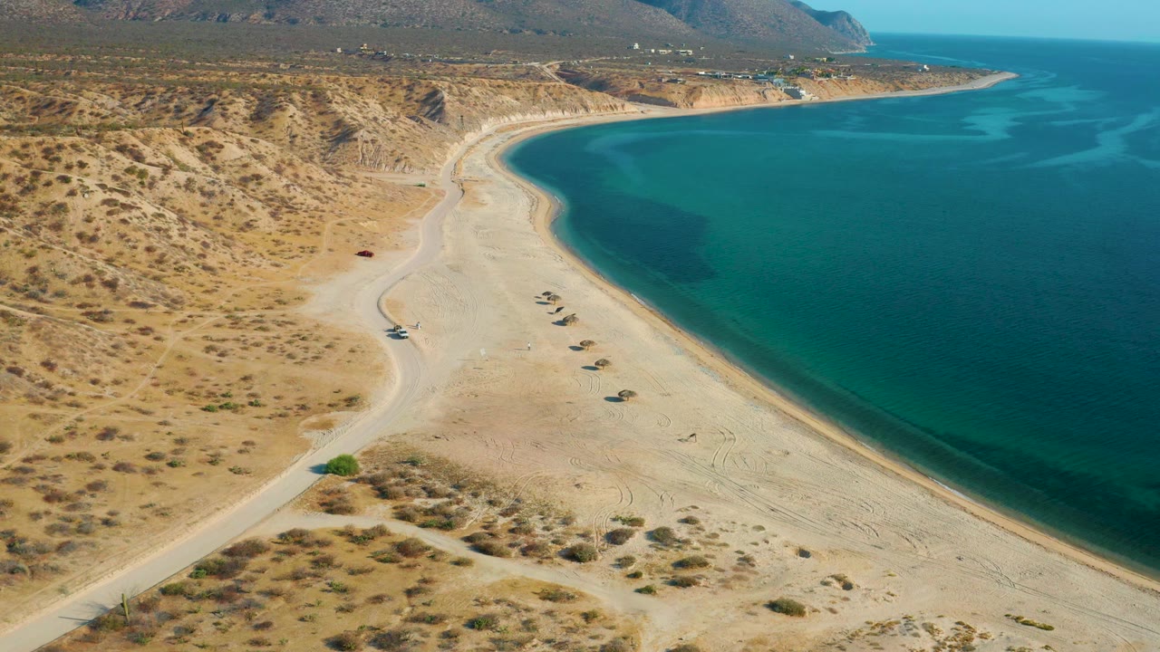 Curved shoreline with pale sand, calm water, and dry hills beyond.