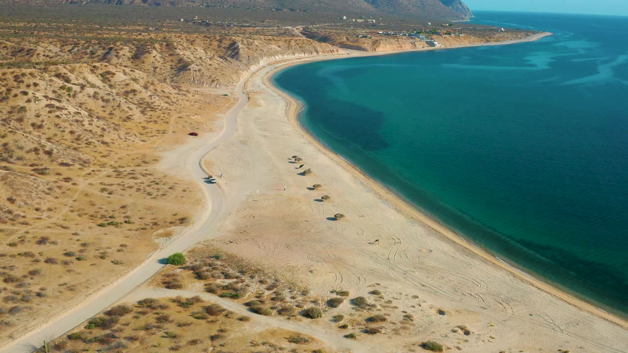 Elevated view over El Sargento La Ventana, Mexico, with turquoise shallows, a broad pale beach, scrub-covered desert terrain, and no visible people.