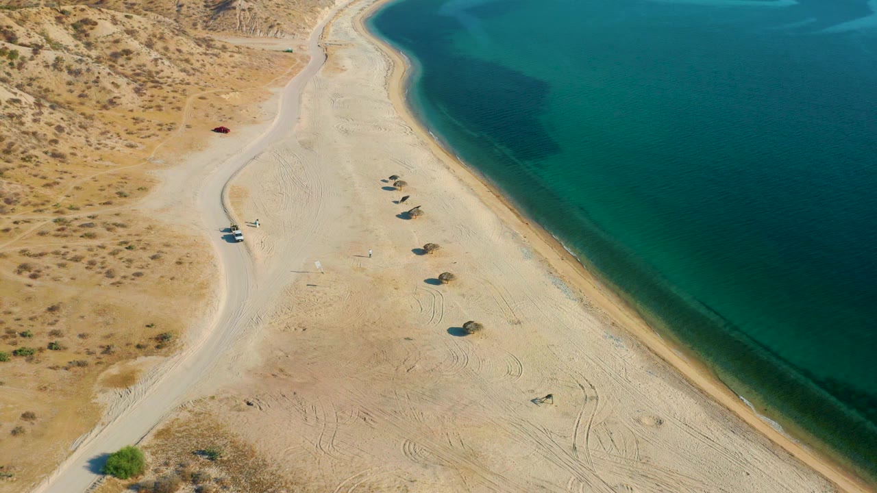 Aerial view in El Sargento La Ventana with the beach centered, desert scrub at upper left, several small shade structures on the sand, and no visible people.