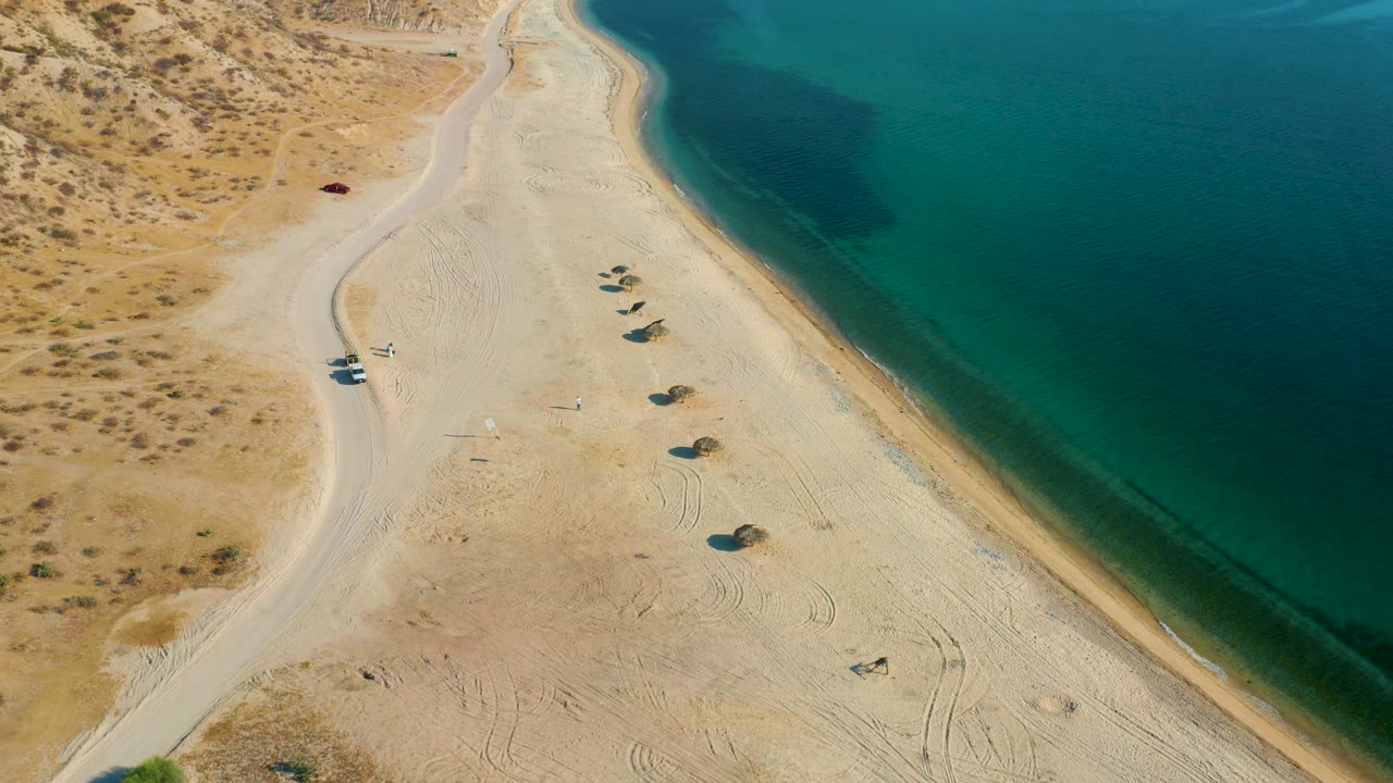 High drone-style frame over El Sargento La Ventana showing a diagonal shoreline, dark green patch in the water, parked vehicles by the road, and no visible people.