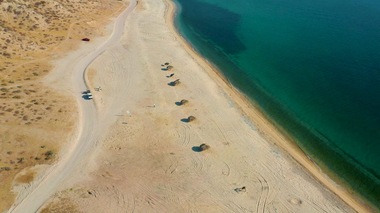 Aerial frame in El Sargento La Ventana with the shoreline running along the right side, tire tracks across the sand, a road on the left, and no visible people.