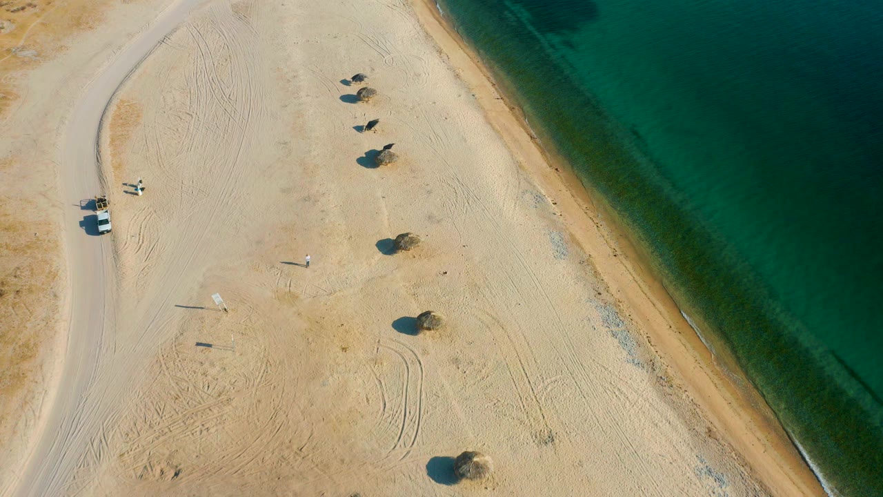 High aerial view of El Sargento La Ventana beach with pale sand, a curved road, scattered small palms, and green-blue water along the right edge, with no visible people.