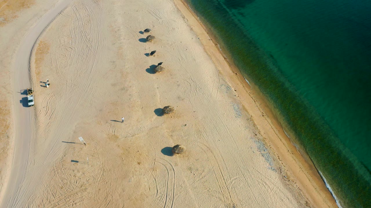 High drone view of the El Sargento La Ventana shoreline with a pale sand beach, a road entering along the left edge, small beach plants, and green water to the right, with no visible people.