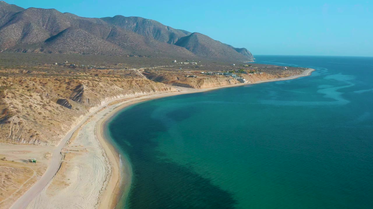 High aerial frame of El Sargento La Ventana coastline showing the pale beach arc, a road along the dry shore, turquoise shallows, distant mountain slopes, and no visible people.