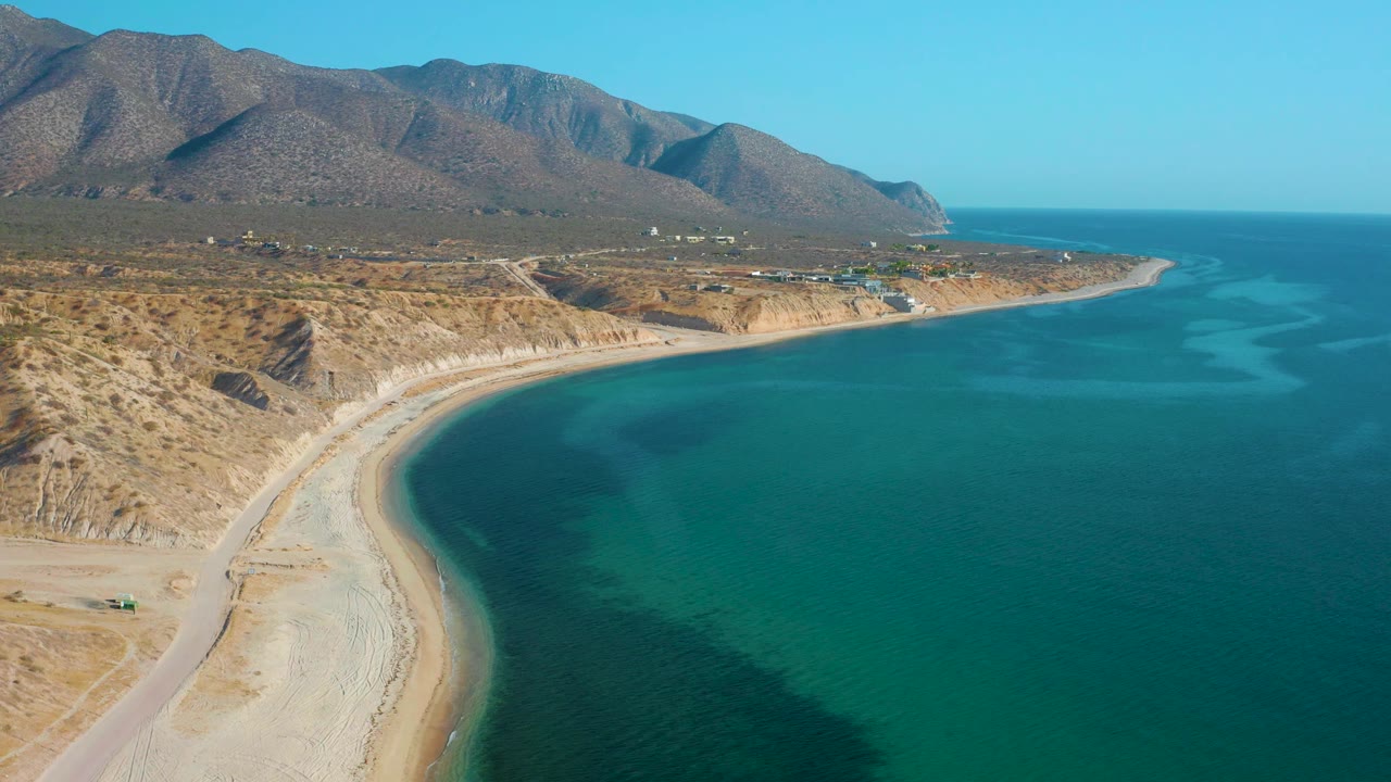 Wide aerial view in El Sargento La Ventana, Mexico, with blue-green water stretching across the right side, a pale curving beach below, dry coastal land, mountain ridges, and no visible people.
