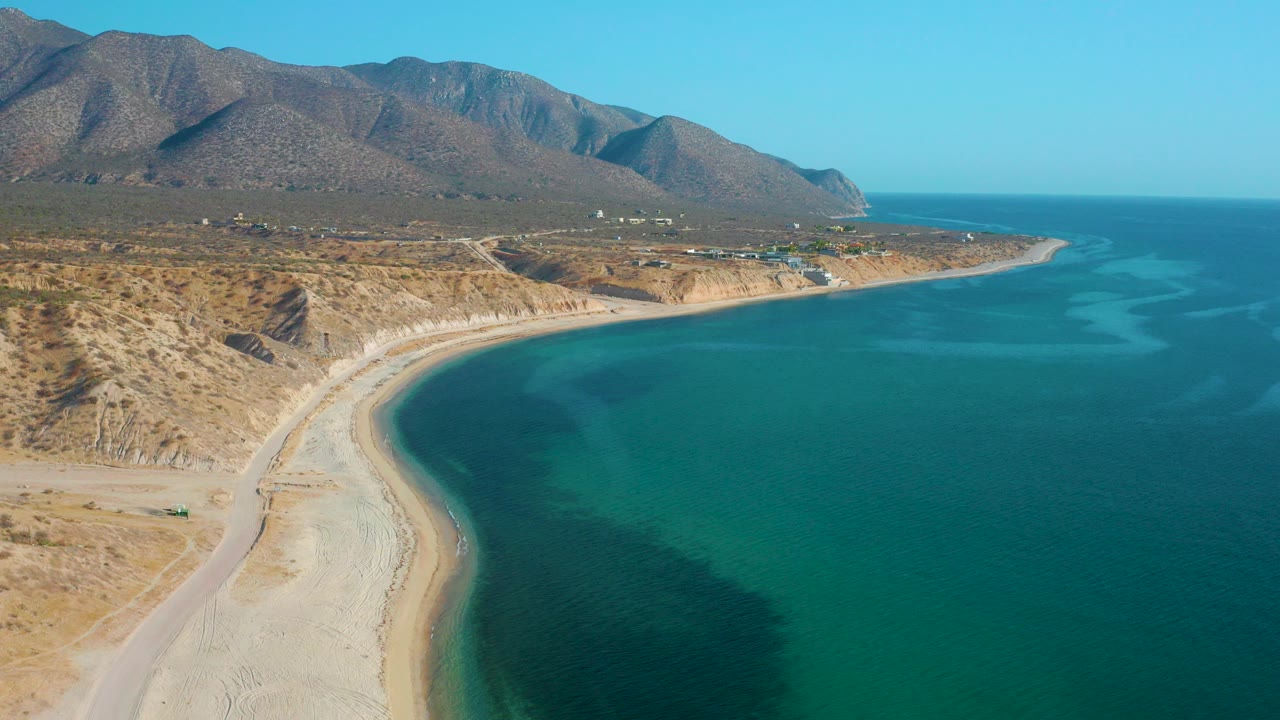 Wide aerial frame over El Sargento La Ventana, Mexico showing a curved pale beach, turquoise water, dirt road, dry hills, rugged mountains, and no visible people.