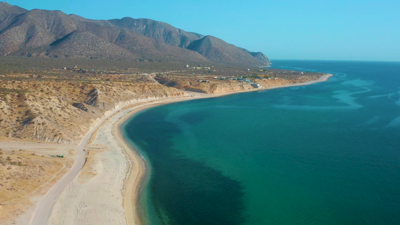 High aerial view in El Sargento La Ventana, Mexico with the empty beach curve more centered, blue-green sea to the right, mountain slopes behind, and no visible people.