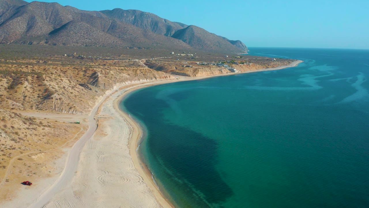 Aerial frame over El Sargento La Ventana, Mexico, with the sandy beach sweeping toward the middle distance, turquoise water on the right, dry mountain slopes behind, and no visible people.