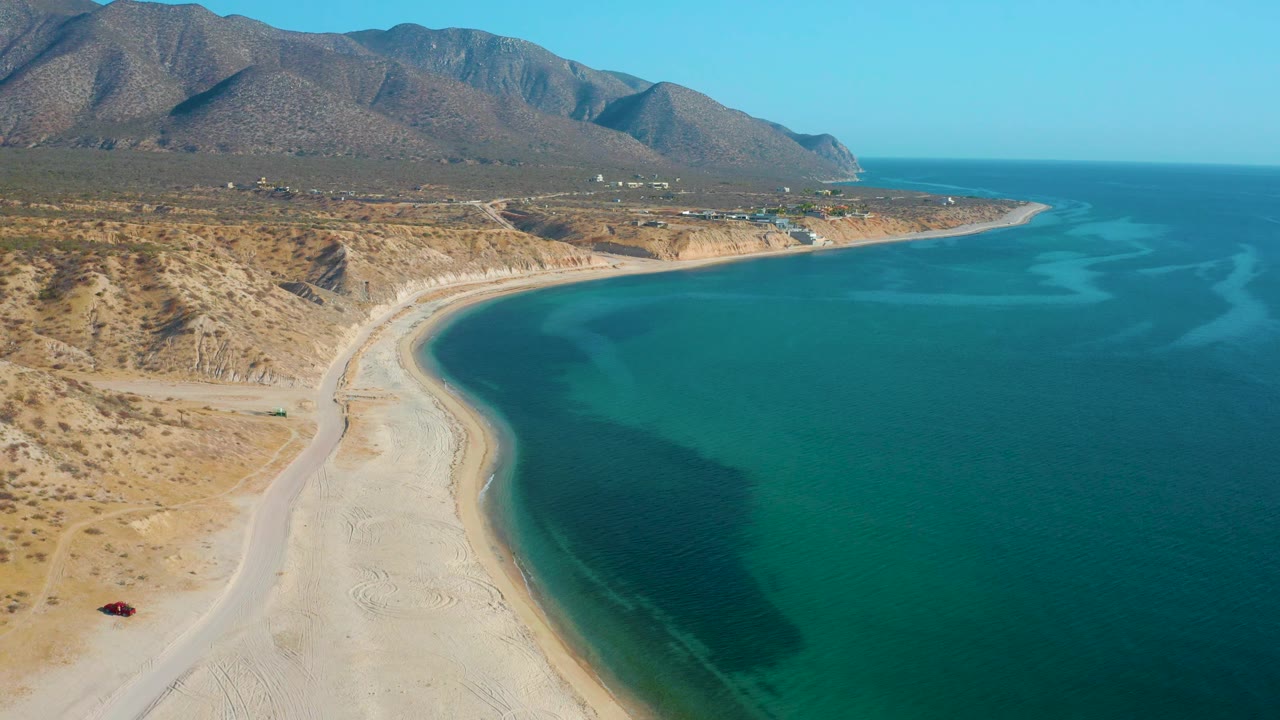 High aerial view in El Sargento La Ventana, Mexico, showing more open ocean to the right of the pale beach, with dry coastal hills and a mountain range under clear light, no visible people.