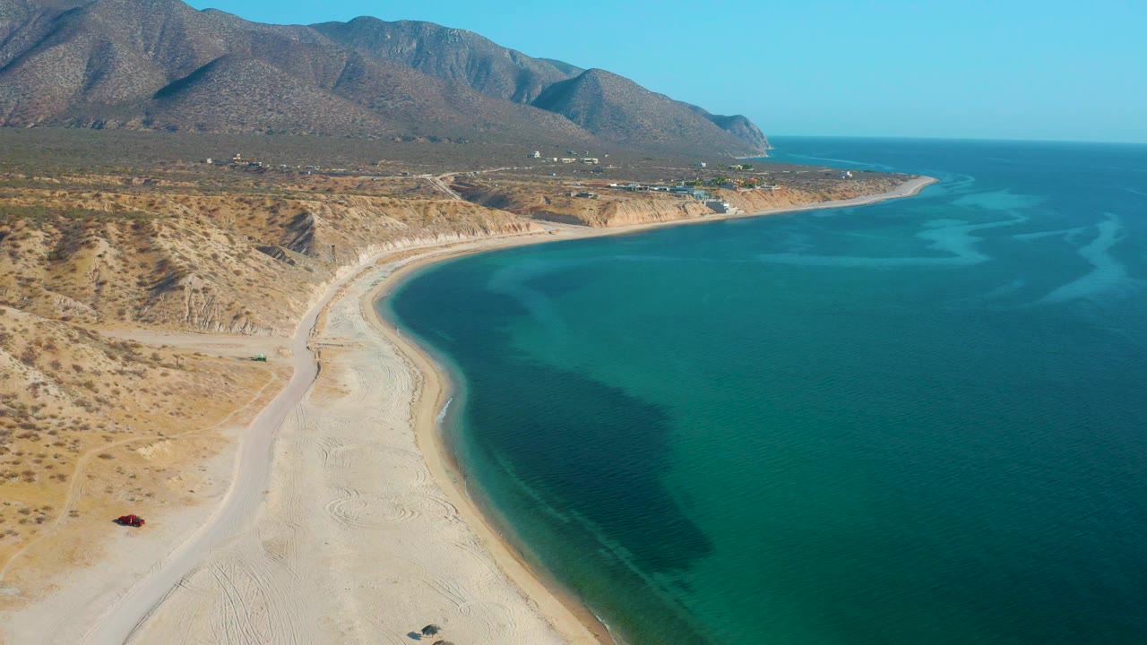 Aerial frame over El Sargento La Ventana, Mexico, where the beach curve sits lower in frame, the ocean fills the right side, dry hills line the left, and no visible people are present.