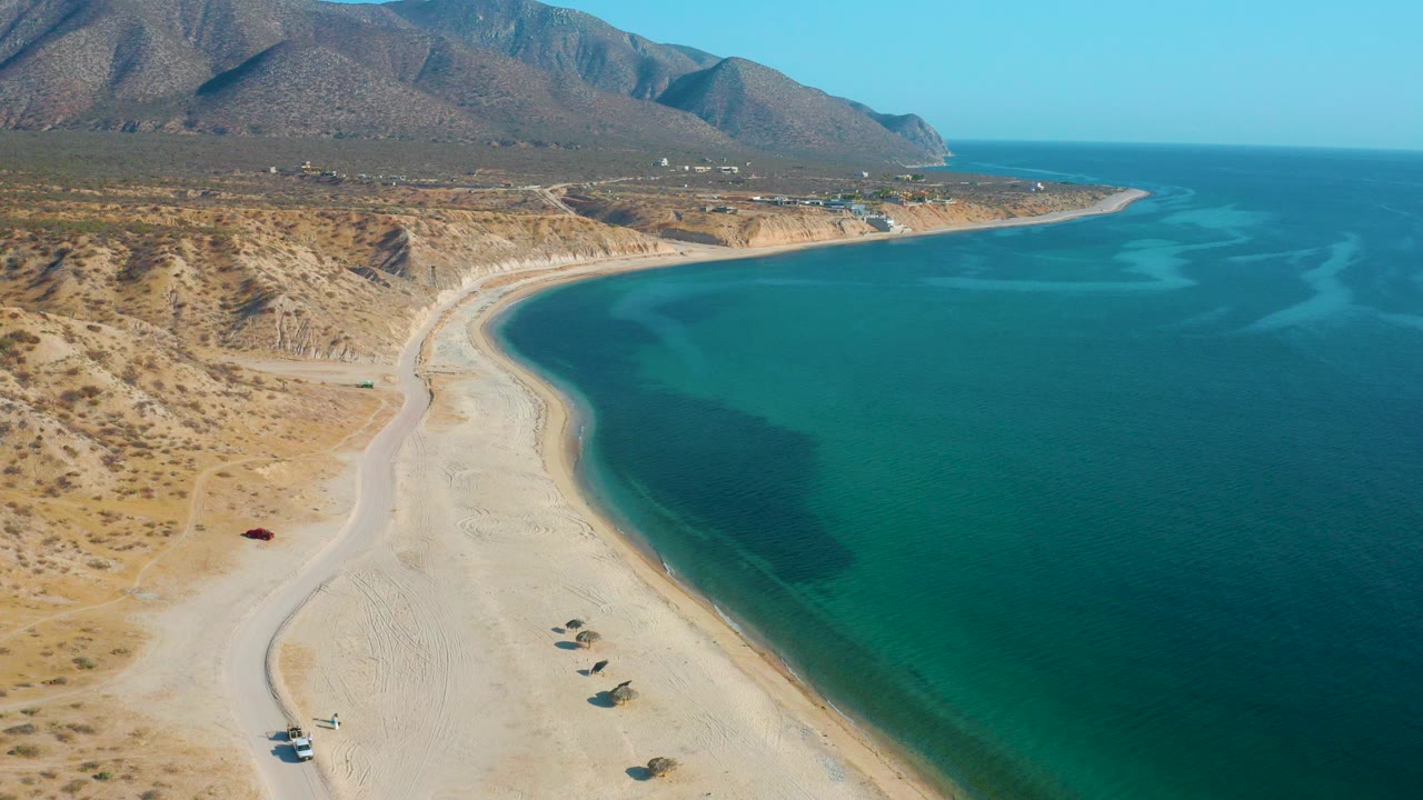 Aerial view of a curved sandy beach beside turquoise water and desert hills.