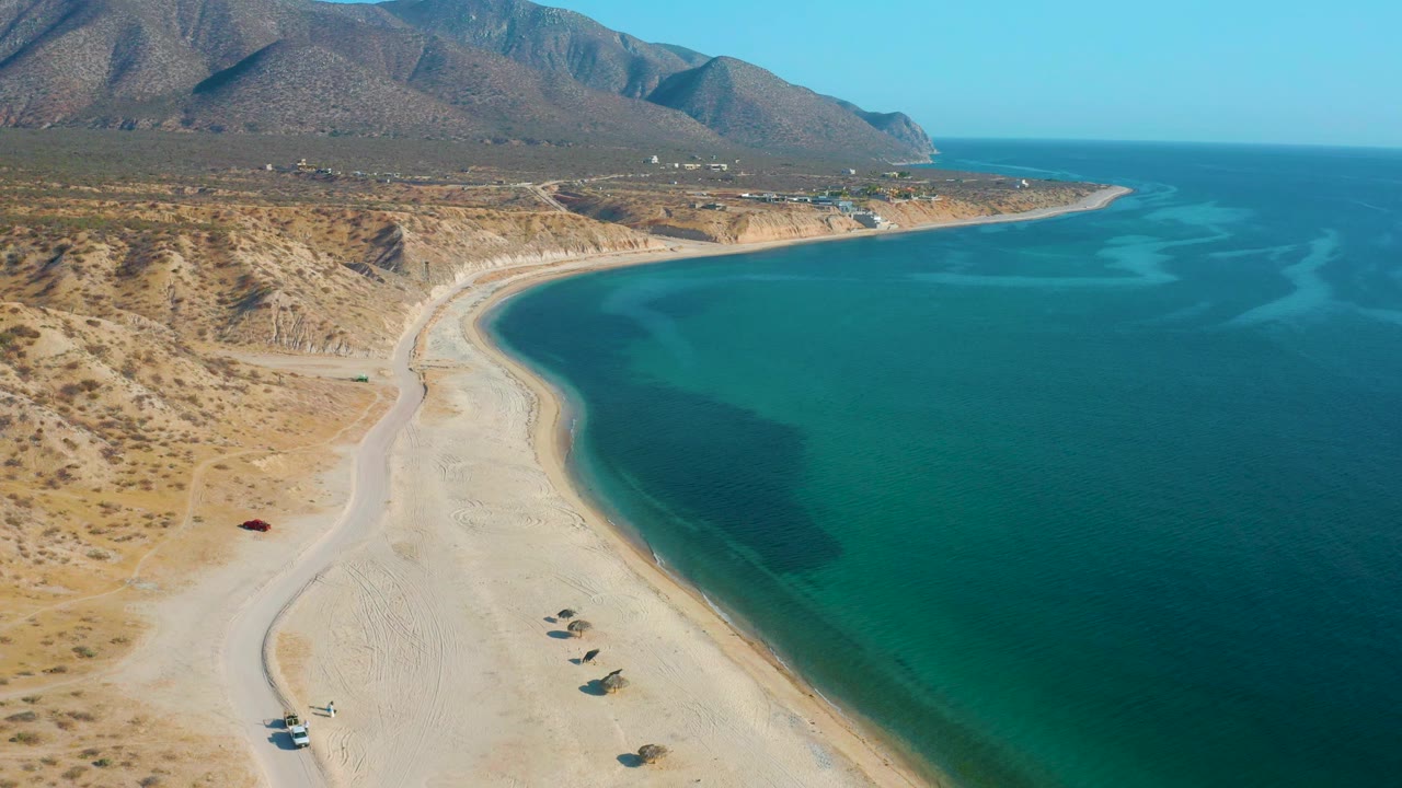 Aerial view over El Sargento La Ventana, Mexico, with the sandbar-like curve of beach leading toward distant hills and no visible people in the frame.