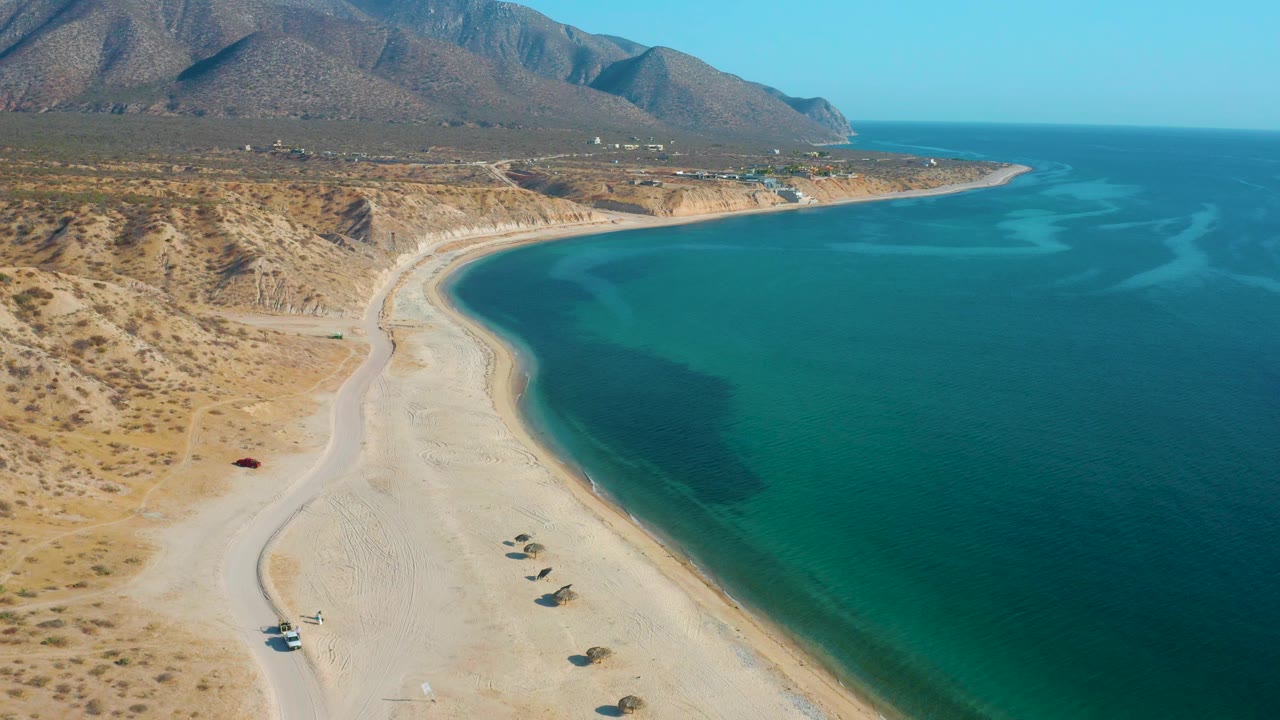 El Sargento La Ventana coastline from above, showing a light beach track, empty sand, turquoise bay water, rugged hills, and no visible people.