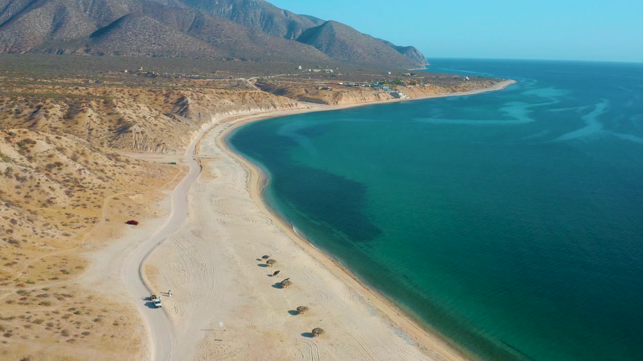 Mid-clip aerial frame over El Sargento La Ventana, Mexico, with desert scrub at left, a pale beach below, blue-green water at right, and no visible people.