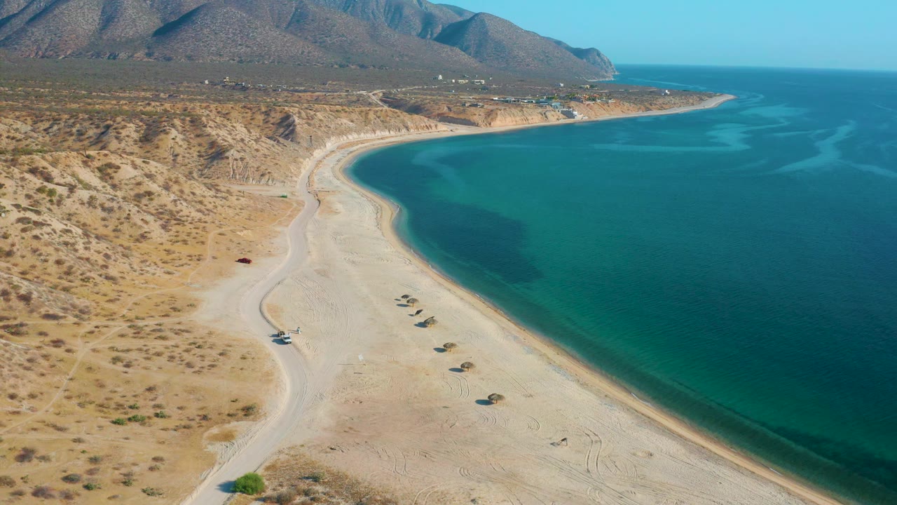 Aerial frame over El Sargento La Ventana with the shoreline bending across the center, deeper blue water at right, dry hills at left, and no visible people.
