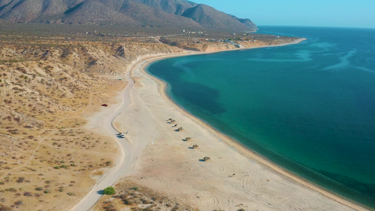 Aerial view in El Sargento La Ventana, Mexico, with empty pale sand, calm blue-green sea, a mountain ridge in the distance, and no visible people.